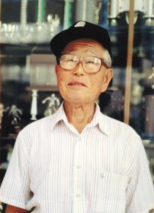 An older Japanese man with large, square glasses, a baseball cap, and a button down short-sleeved shirt with thin pastel stripes. He smiles gently for the camera, standing in front of a trophy shop window. From the film 95 and 6 to Go.